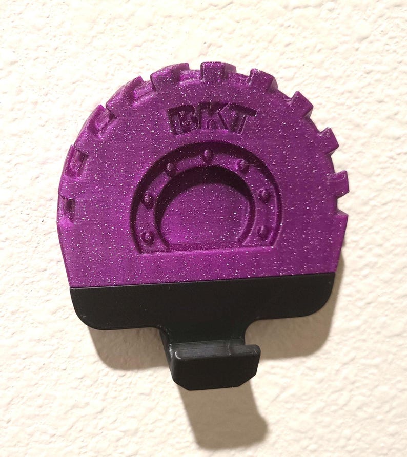 May include: A purple, glittery, gear-shaped object with the letters "BKT" embossed on the top. The object has a black base with a small, rectangular protrusion. The object is likely a decorative or functional item.