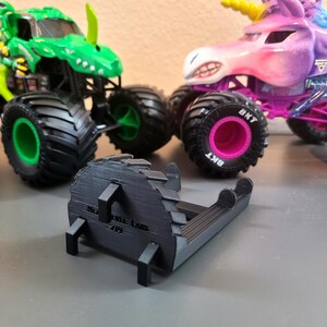 May include: A black 3D-printed object with the text "NEW WHEEL LABS 719" is in the foreground. Two toy monster trucks are in the background, one green and one purple with a unicorn design. The trucks have large black tires.