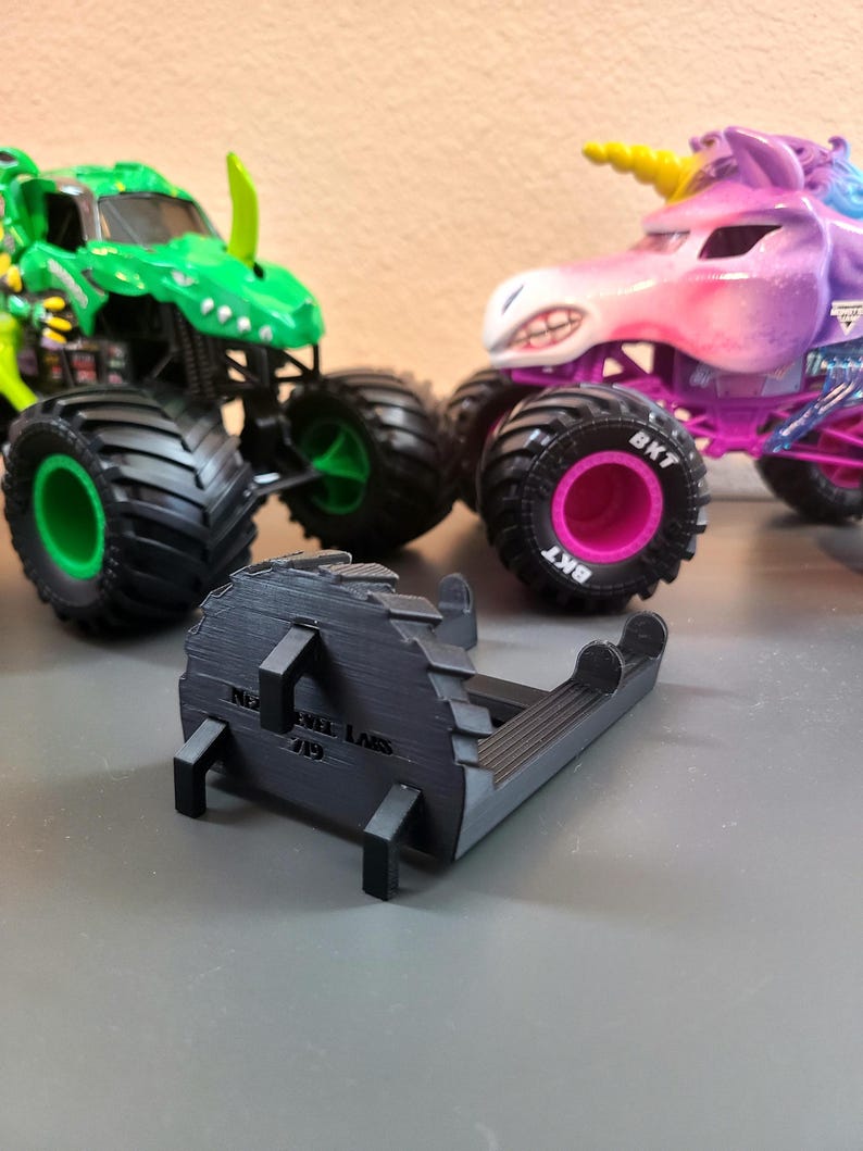 May include: A black 3D-printed object with the text "NEW WHEEL LABS 719" is in the foreground. Two toy monster trucks are in the background, one green and one purple with a unicorn design. The trucks have large black tires.