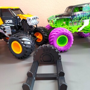 May include: Image of two toy monster trucks, one black and yellow, the other green and black, with large tires. A black tire stand with the text "BKT" is in the foreground. The trucks have orange, purple, and green accents.