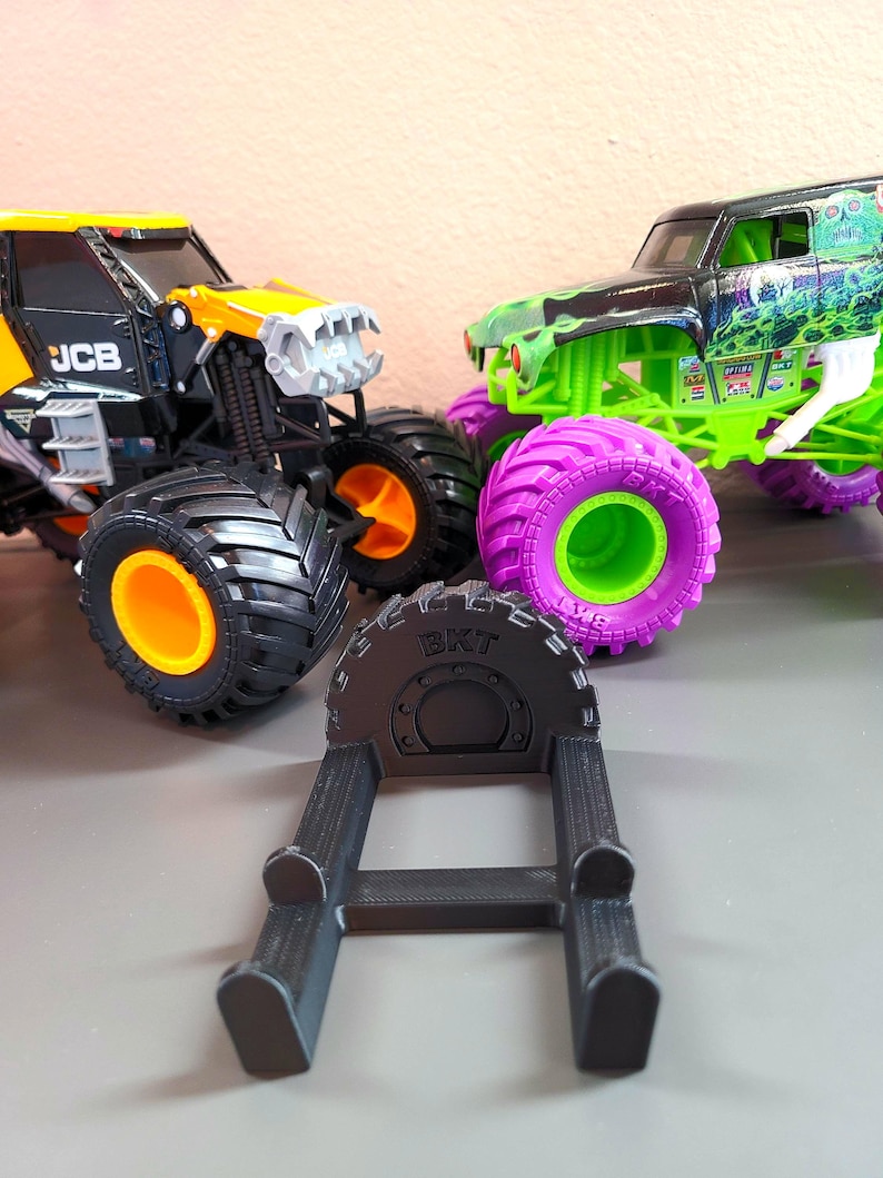 May include: Image of two toy monster trucks, one black and yellow, the other green and black, with large tires. A black tire stand with the text "BKT" is in the foreground. The trucks have orange, purple, and green accents.