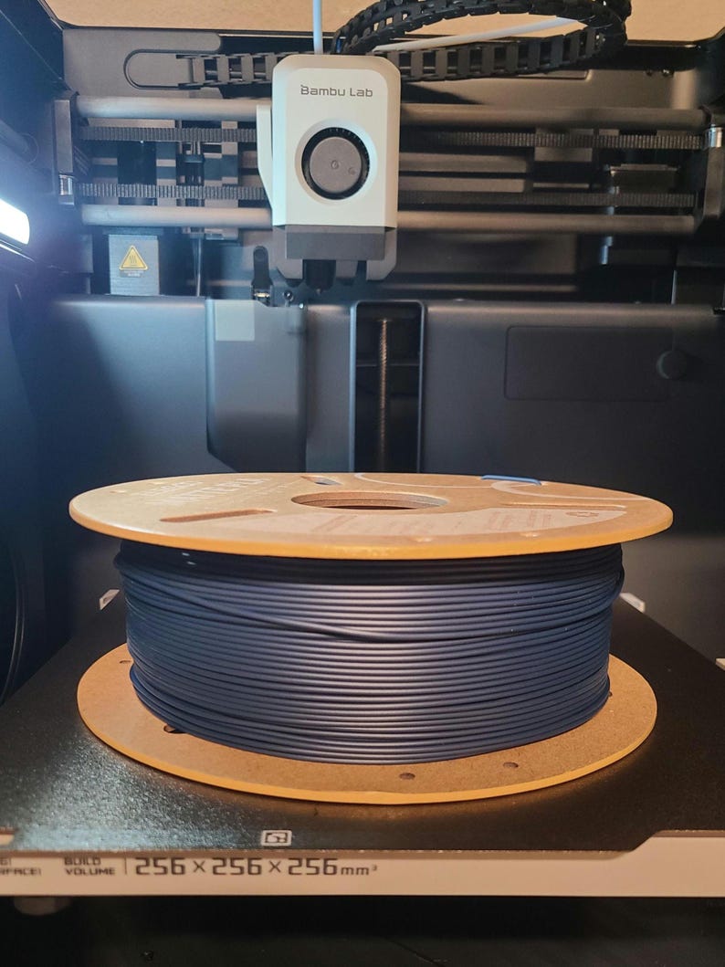May include: A spool of dark blue filament sits inside a 3D printer. The printer has the text "Bambu Lab" on the print head. The build volume is 256 x 256 x 256 mm.