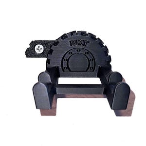 May include: A black, 3D-printed wall mount with the letters "BKT" on the top. The mount has a gear-like design and two curved arms extending from a horizontal bar. A small plate with a screw is attached to the side.