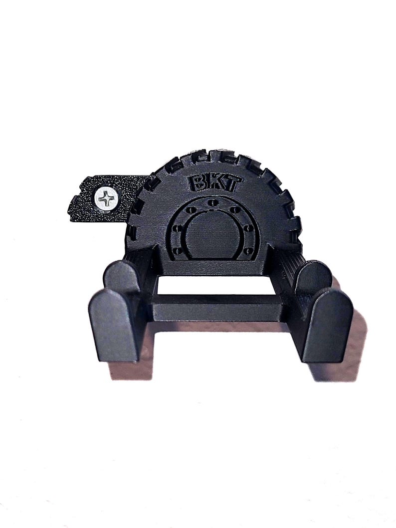 May include: A black, 3D-printed wall mount with the letters "BKT" on the top. The mount has a gear-like design and two curved arms extending from a horizontal bar. A small plate with a screw is attached to the side.