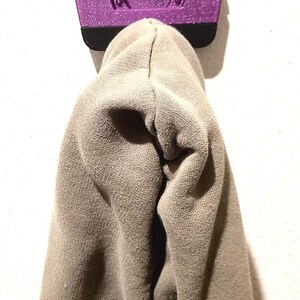 May include: A purple wall hook with the letters "BKT" on top, holding a tan hoodie. The hook has a textured, glittery finish and a black base. The hoodie is made of a soft, knit fabric.