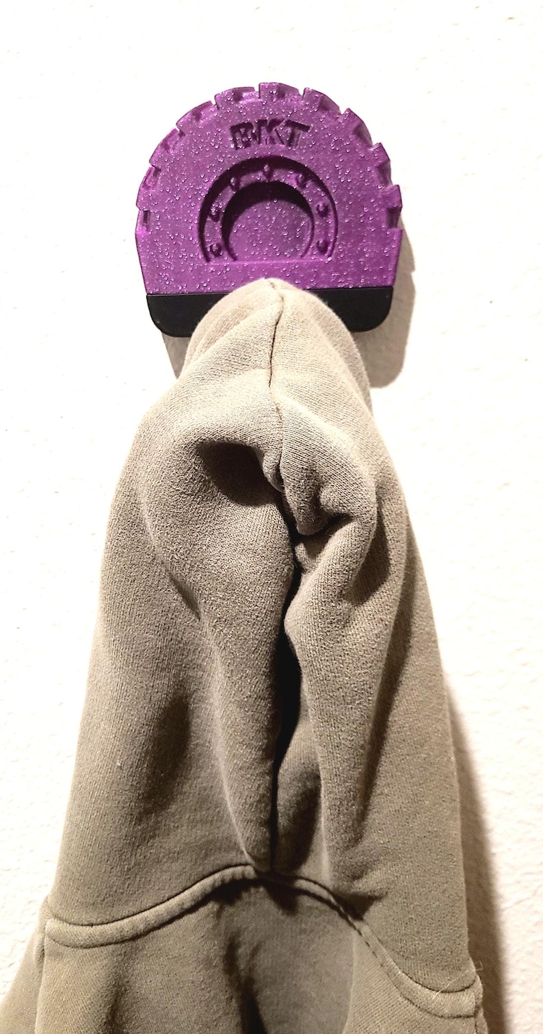May include: A purple wall hook with the letters "BKT" on top, holding a tan hoodie. The hook has a textured, glittery finish and a black base. The hoodie is made of a soft, knit fabric.