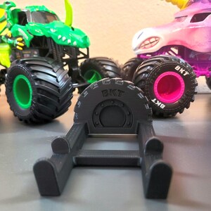 May include: A black 3D-printed tire stand with the text "BKT" on the tire. In the background are two toy monster trucks: a green dinosaur truck and a pink unicorn truck. The monster trucks have large black tires.