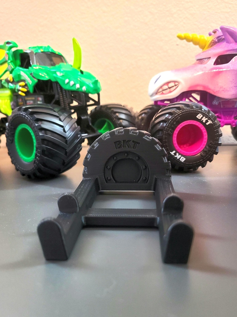 May include: A black 3D-printed tire stand with the text "BKT" on the tire. In the background are two toy monster trucks: a green dinosaur truck and a pink unicorn truck. The monster trucks have large black tires.
