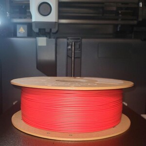May include: A red filament spool sits inside a 3D printer. The spool is made of a bright red plastic and is mounted on a wooden base. The printer has a white and black print head and a build volume of 256 x 256 x 256 mm.