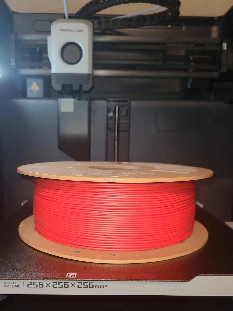 May include: A red filament spool sits inside a 3D printer. The spool is made of a bright red plastic and is mounted on a wooden base. The printer has a white and black print head and a build volume of 256 x 256 x 256 mm.