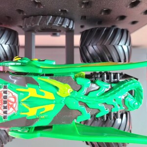 May include: Green and gray toy monster truck with large black tires. The truck features a detailed design with the word "BAKUGAN" visible on the side. The truck is designed with a green and yellow color scheme.