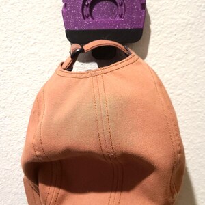 May include: A peach-colored baseball cap hanging from a purple wall hook. The hook has the letters "BKT" on it. The cap is made of a soft material and has a curved brim.