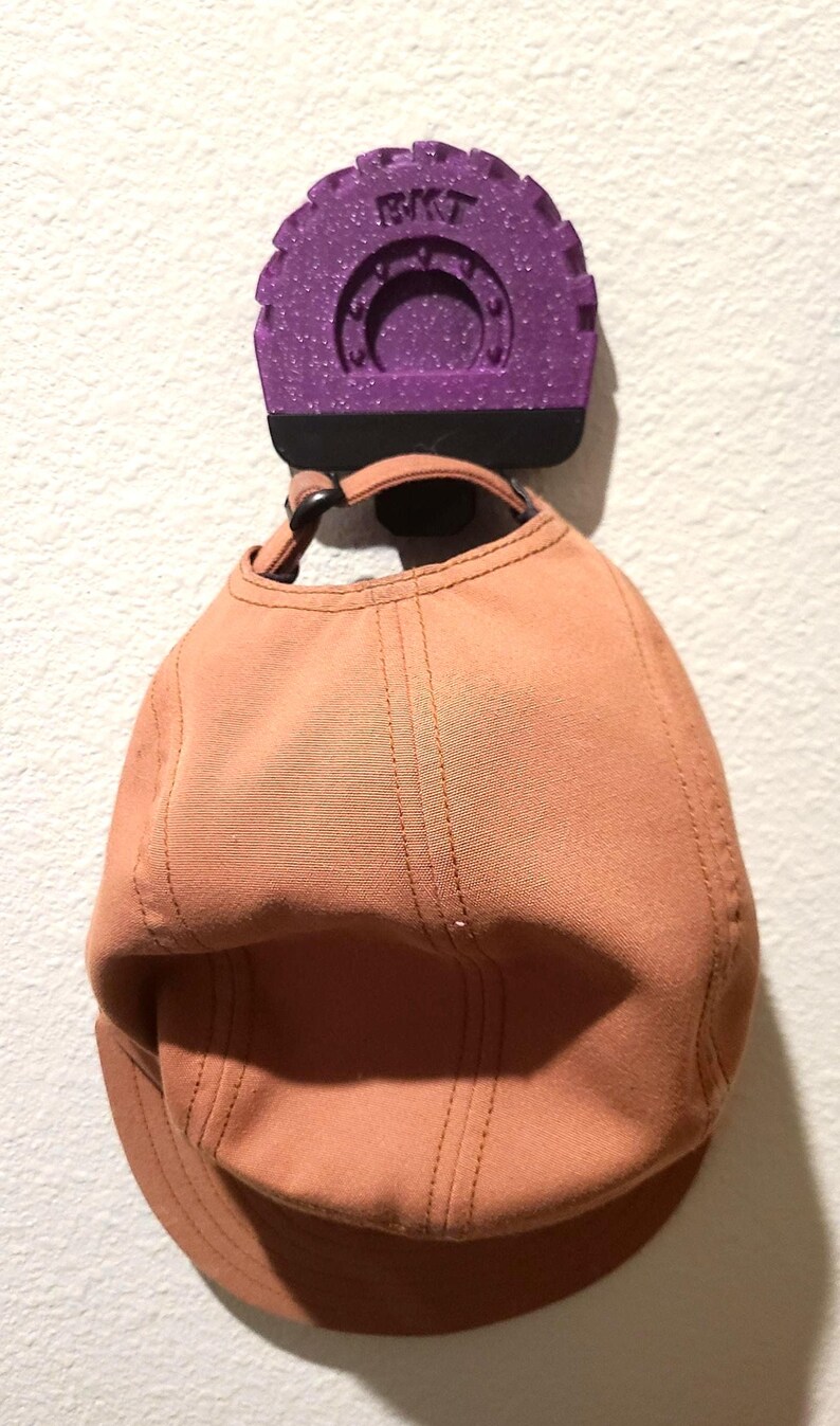 May include: A peach-colored baseball cap hanging from a purple wall hook. The hook has the letters "BKT" on it. The cap is made of a soft material and has a curved brim.