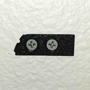 May include: A black, textured metal plate with two silver screws. The plate has a unique, asymmetrical shape and is mounted on a white, textured wall. The screws are evenly spaced and have a cross-shaped indentation.