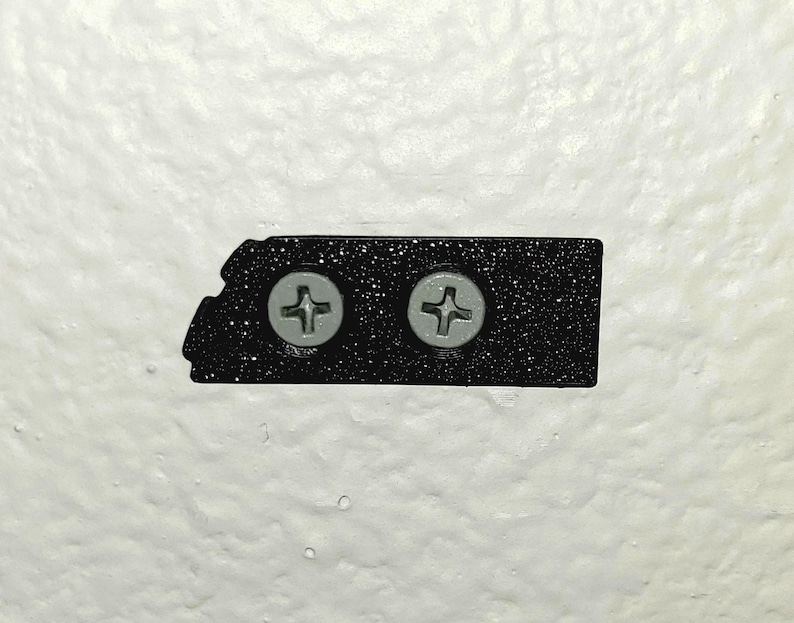 May include: A black, textured metal plate with two silver screws. The plate has a unique, asymmetrical shape and is mounted on a white, textured wall. The screws are evenly spaced and have a cross-shaped indentation.