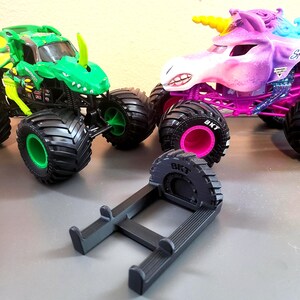 May include: A green monster truck with a dinosaur design and a purple unicorn monster truck with a yellow horn. A black stand is in the foreground. The monster trucks have large black tires with pink and green accents.