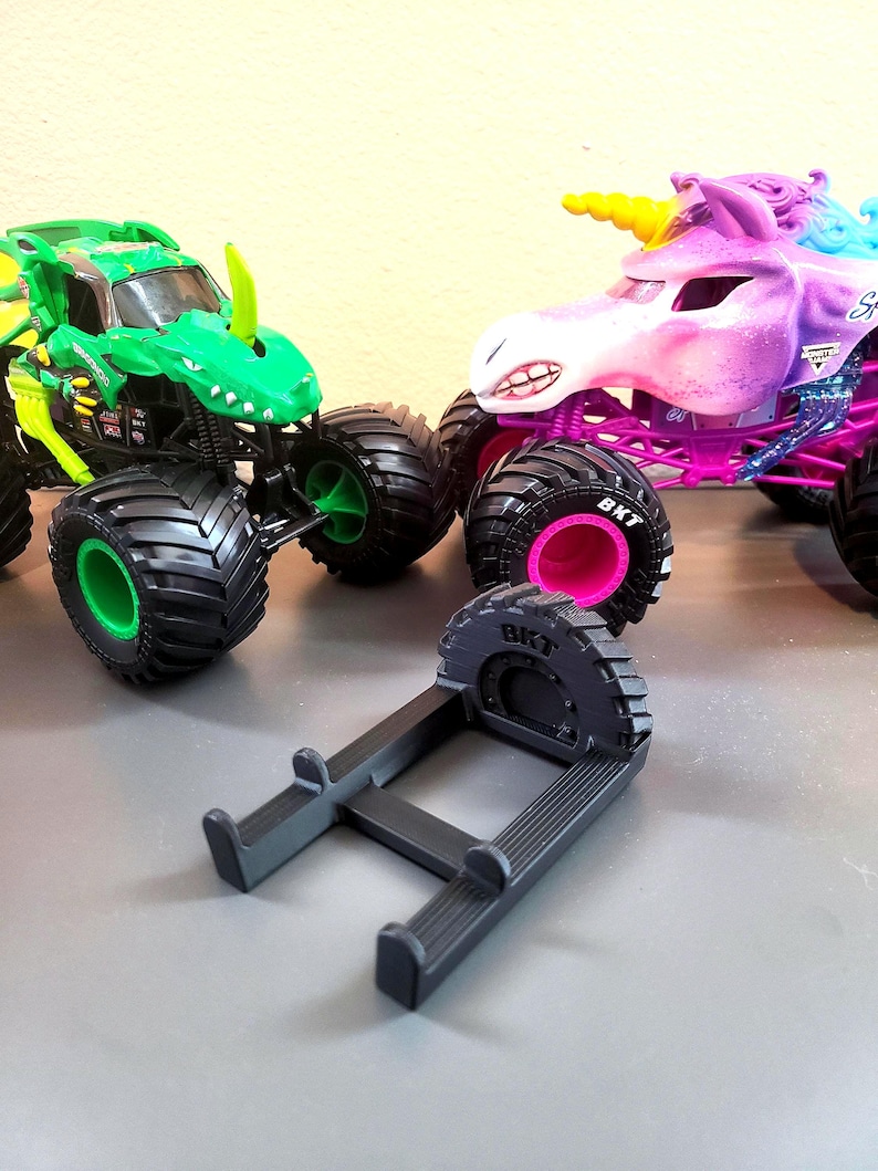 May include: A green monster truck with a dinosaur design and a purple unicorn monster truck with a yellow horn. A black stand is in the foreground. The monster trucks have large black tires with pink and green accents.