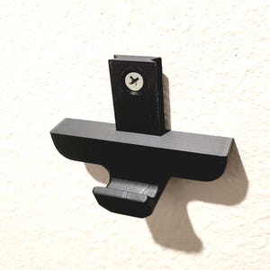 May include: A black, wall-mounted bracket with a cross-shaped design. The bracket is made of a matte material and is attached to the wall with a single screw. The design includes a hook-like feature on the bottom.