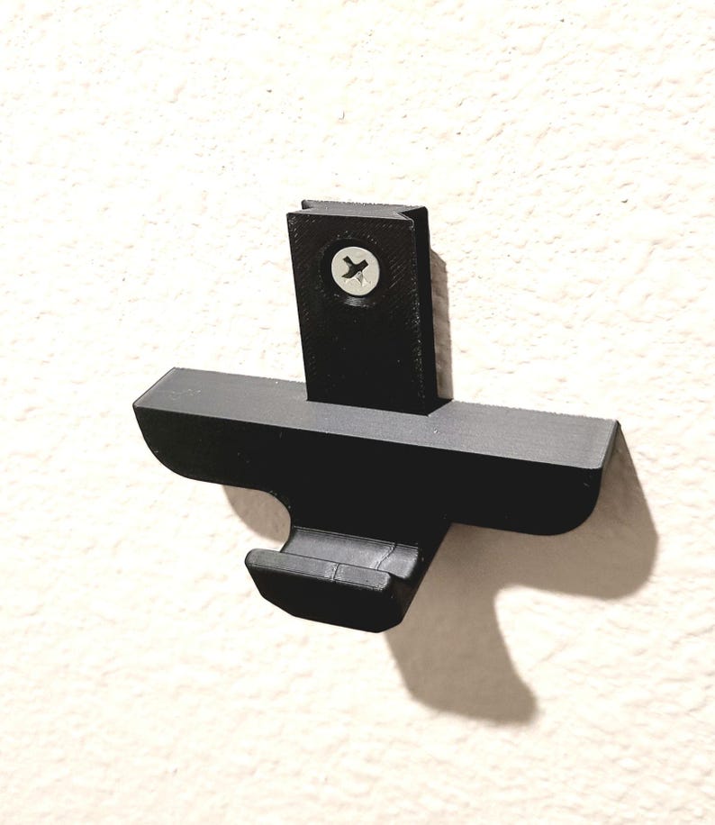 May include: A black, wall-mounted bracket with a cross-shaped design. The bracket is made of a matte material and is attached to the wall with a single screw. The design includes a hook-like feature on the bottom.