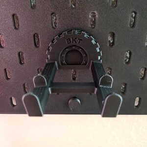 May include: A black pegboard accessory with the letters "BKT" on a circular gear-like component. The accessory has two arms extending downwards, designed to hold items. The pegboard itself is black with evenly spaced oval holes.