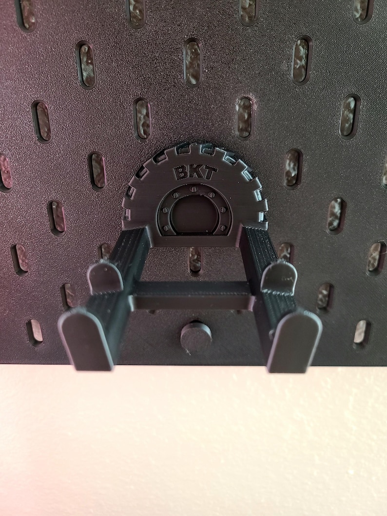 May include: A black pegboard accessory with the letters "BKT" on a circular gear-like component. The accessory has two arms extending downwards, designed to hold items. The pegboard itself is black with evenly spaced oval holes.