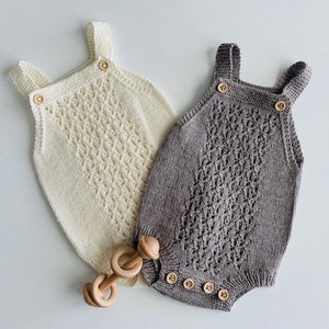 May include: Two knitted baby onesies with straps, one in white and one in gray. Both onesies have a delicate lace pattern and wooden buttons.