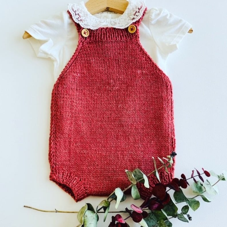 K&ouml;nnte beinhalten: Ein gestrickter Baby-Strampler in tiefroter Farbe mit Holzkn&ouml;pfen. Der Strampler hat einen wei&szlig;en Spitzenkragen und ist auf wei&szlig;em Hintergrund fotografiert.