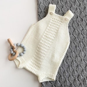 Könnte beinhalten: Ein weißer gestrickter Baby-Strampler mit Trägern und Knöpfen. Der Strampler liegt auf einer grau-weißen Decke.