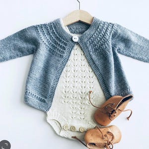 Può includere: Un cardigan in maglia azzurro con chiusura a bottone bianco singolo sovrapposto a un body in maglia color crema con motivo testurizzato e tre bottoni sul fondo, accompagnato da un paio di scarpine da neonato stringate in cuoio.