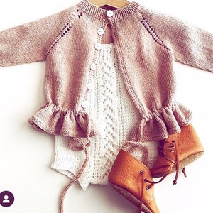 以下が含まれることがあります： 白いボタンとフリルのある裾が付いたピンクのニットカーディガン。カーディガンは、レース模様の白いニットロンパースの上に着用されています。茶色の革製のベビーブーツも写っています。