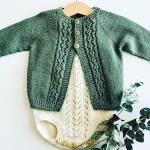Könnte beinhalten: Ein grüner Strickcardigan mit Zopfmuster und Knopfleiste. Der Cardigan wird über einem weißen Strickbody mit Spitzenmuster getragen.