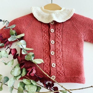 以下が含まれることがあります： 白いクロシェの襟とボタン留めが付いた赤いニットカーディガン。カーディガンはケーブルニットのパターンで、ユーカリの枝と一緒に撮影されています。