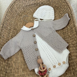 Puede incluir: Un conjunto de ropa de bebé de punto en beige y blanco, que incluye un cárdigan, un mono y un gorro. El conjunto está colocado en una cesta tejida.