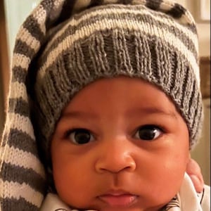 Peut inclure: Gros plan d'un bébé portant un bonnet en tricot à rayures grises et blanches avec une longue extrémité pointue. Le bébé a les yeux sombres et porte un body blanc avec un petit motif.
