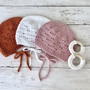 May include: Three knitted baby hats with ties in orange, white, and pink. The hats have a lacy pattern and are displayed on a white wooden surface with two white wooden teething rings.