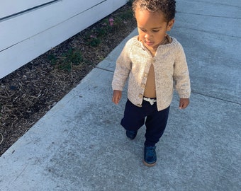 Puede incluir: Un niño pequeño lleva un cárdigan de punto beige con un botón marrón, pantalones azul oscuro y zapatillas azules caminando por una acera de hormigón.