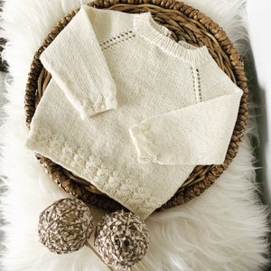 Peut inclure: Un pull en tricot crème pour bébé avec un délicat motif de dentelle autour du col et des manches. Le pull est posé sur une surface en fausse fourrure blanche.