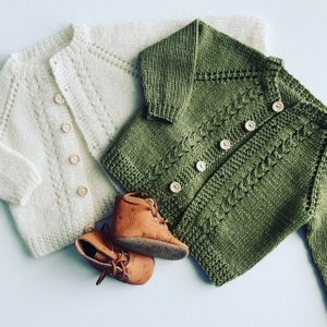 May include: Two knitted baby cardigans, one white and one green, with button closures. The cardigans are laid out on a white surface with two pairs of brown leather baby shoes.