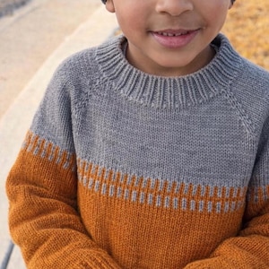 May include: A grey and orange knitted jumper with a matching grey beanie. The jumper has a ribbed collar and cuffs, with an orange band across the chest and sleeves. The child is smiling.