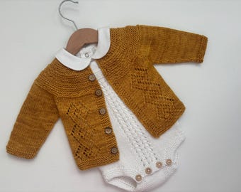 Könnte beinhalten: Ein gestrickter Baby-Cardigan in senfgelber Farbe mit einem weißen, gehäkelten Strampler darunter. Der Cardigan hat einen weißen Kragen und einen Knopfverschluss.