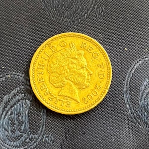 May include: A gold-colored coin with the profile of a person and the text "ELIZABETH II D G REG FD 2002". The coin has a textured edge and is set against a dark background.