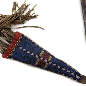 May include: A beaded sheath with a brown leather fringe and a red and gold trim. The sheath is primarily blue with a geometric pattern in red, brown, and gold. A wooden stick is visible in the background.