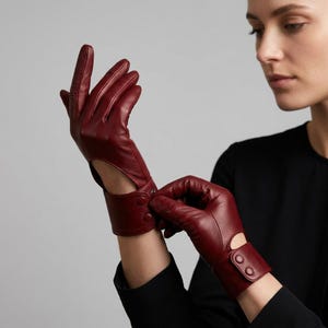 May include: A pair of deep red leather gloves featuring a wrist-length design with a cutout detail on the back of the hand. Each glove includes a wide cuff secured by two matching red snap buttons, providing a structured fit around the wrist area.