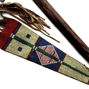 May include: A beaded sheath with a red top and a geometric design in blue, green, and pink. The sheath is adorned with leather fringe and lies next to a wooden handle, likely for a knife or similar tool.
