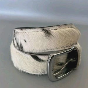 Op de afbeelding: Een riem met een gestructureerd wit en zwart dierenharen patroon. De riem is opgerold en bevat een gepolijste zilverkleurige metalen gesp met een rechthoekig frame en een enkele doorn, met een natuurlijke haar-op-huid afwerking.