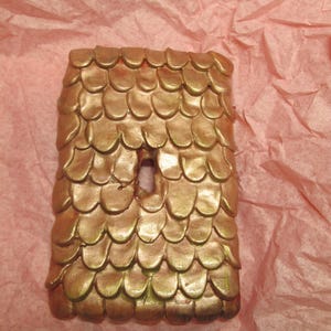 Puede incluir: Una tapa de interruptor de luz de cerámica de color dorado con un diseño texturizado en forma de escamas. La tapa tiene una cerradura en el centro.