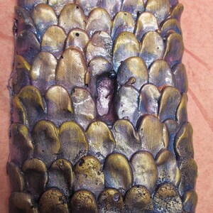 Puede incluir: Una pieza de arte rectangular y texturizada con un patrón similar a escamas. Las escamas son una mezcla de tonos metálicos dorados, bronce y morados, creando un efecto dimensional. La pieza tiene una apariencia áspera y artesanal.