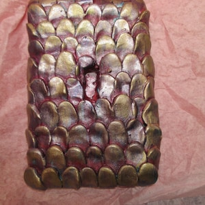 Puede incluir: Objeto rectangular con un patrón similar a escamas. Las escamas son una mezcla de dorado y burdeos, con toques de azul. El objeto parece hecho a mano, con una superficie texturizada y una forma ligeramente irregular. El fondo es rosa claro.