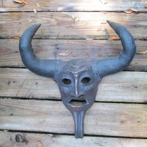 May include: A dark gray bull mask with large curved horns, a carved face, and a long, pointed chin. The mask is made of a dark material and rests on a wooden surface.