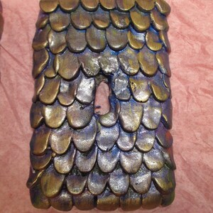 Puede incluir: Un objeto rectangular hecho a mano con un ojo de cerradura, cubierto de formas superpuestas en forma de escamas. Las escamas son principalmente de bronce y oro, con toques de azul y morado. El objeto tiene un aspecto texturizado y metálico.